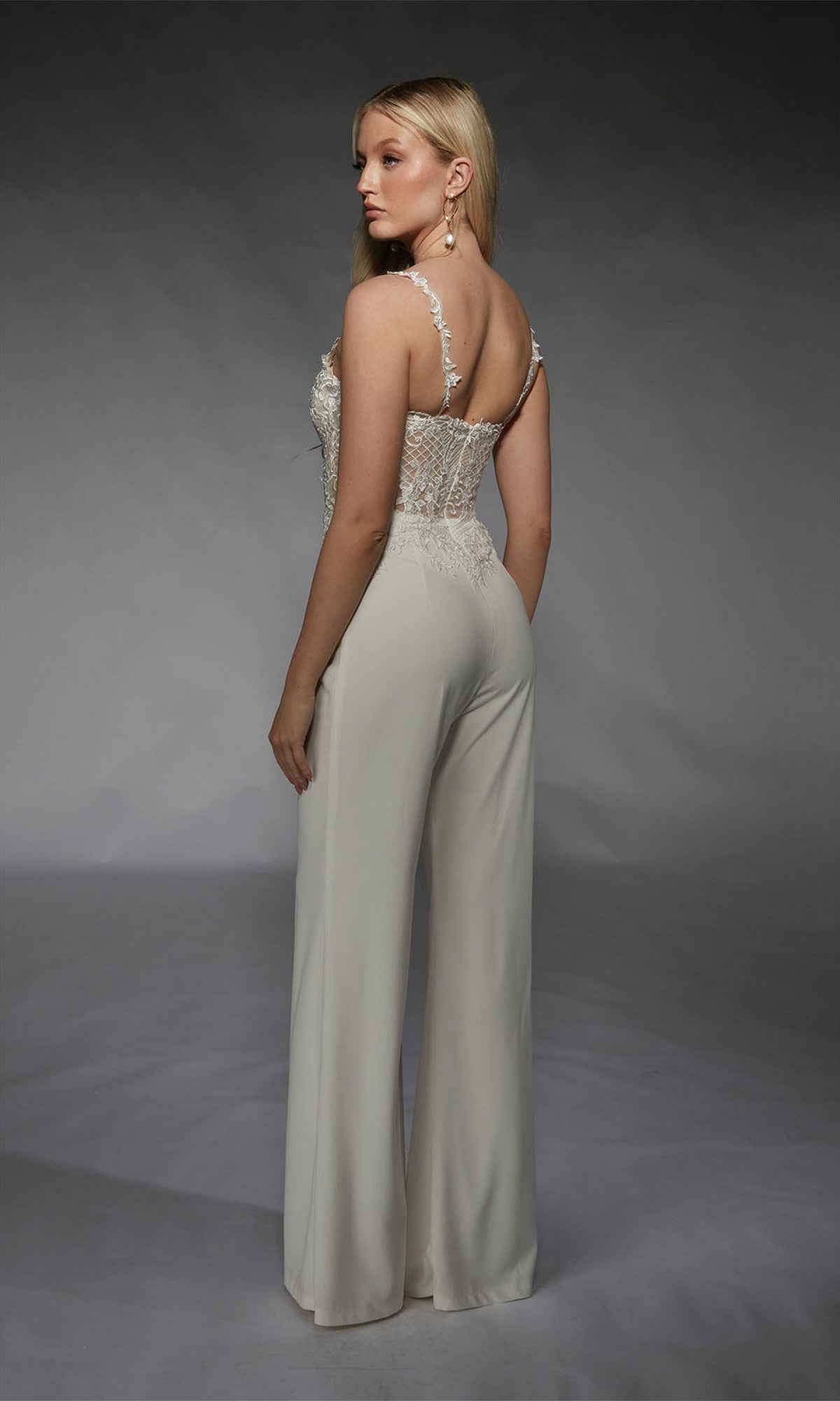 A woman models the Alyce 70074 long formal jumpsuit, showcasing a sleeveless white lace corset top and high-waisted wide-leg pants, set against a plain studio background.